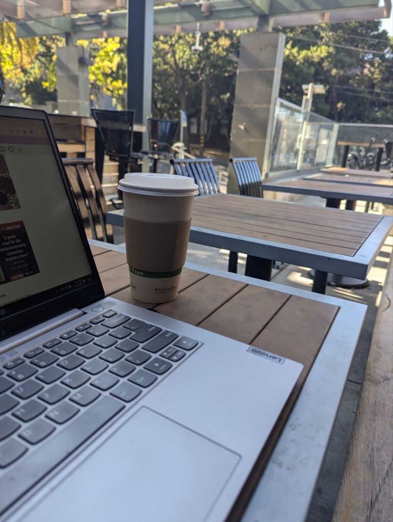 To go cup of Cafe au lait and laptop on a table on an outside coffee shop patio because I am starting work outside today 