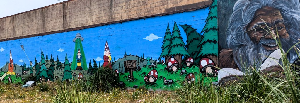 Picture of a mural on a house wall. There is gnomes in the forest (one taken away by an alien ship) and on the right a face of a man smiling.