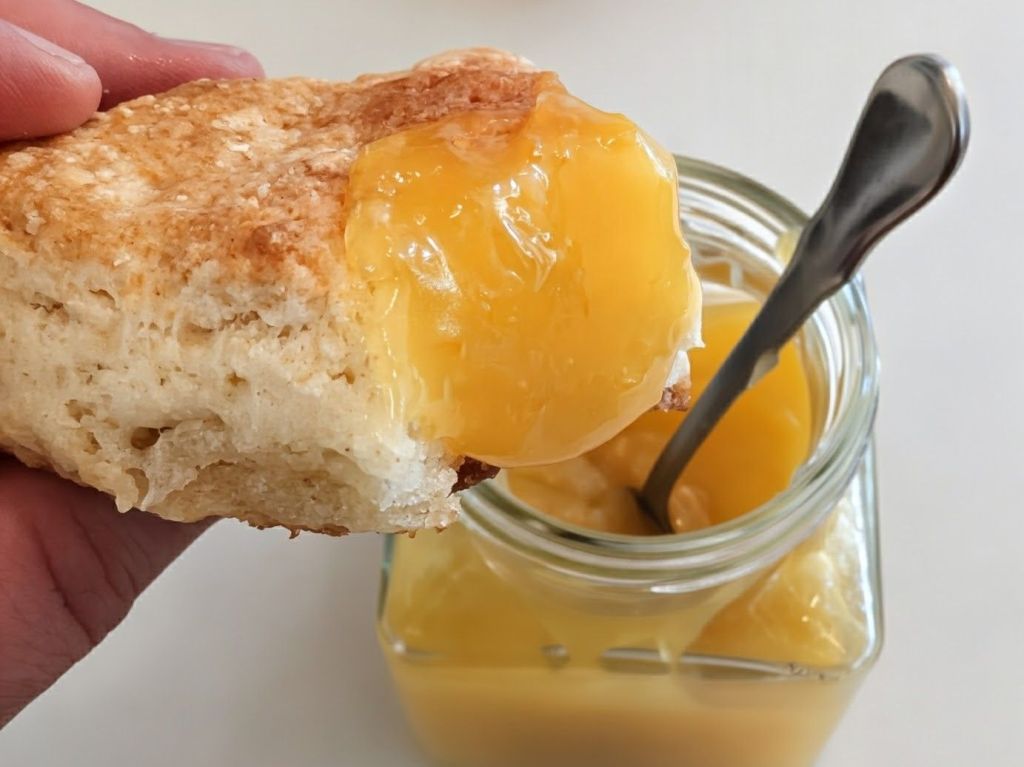 Someone holding a scone with a dollop of lemon curd, next to a glass of lemon curd with a spoon inside.
